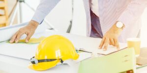 A group of people are gathered around a table, looking at blueprints and a yellow hard hat.