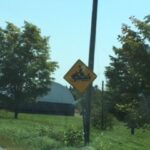 Roadside caution sign with a tractor symbol, indicating a farming or tractor crossing area, surrounded by trees and grass on a sunny day.