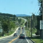 A straight, two-lane road with trees and hills in the distance.